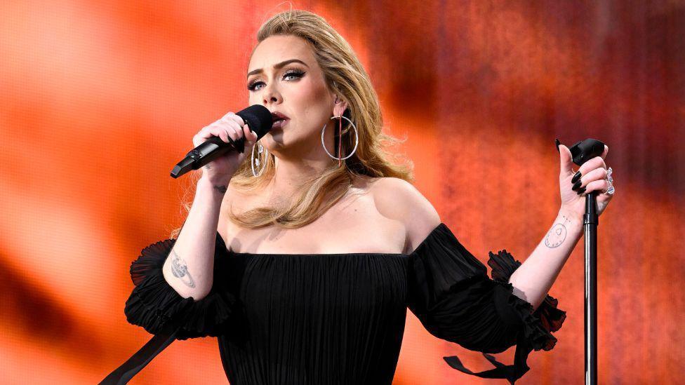 Adele wearing a black dress and singing into a microphone. 