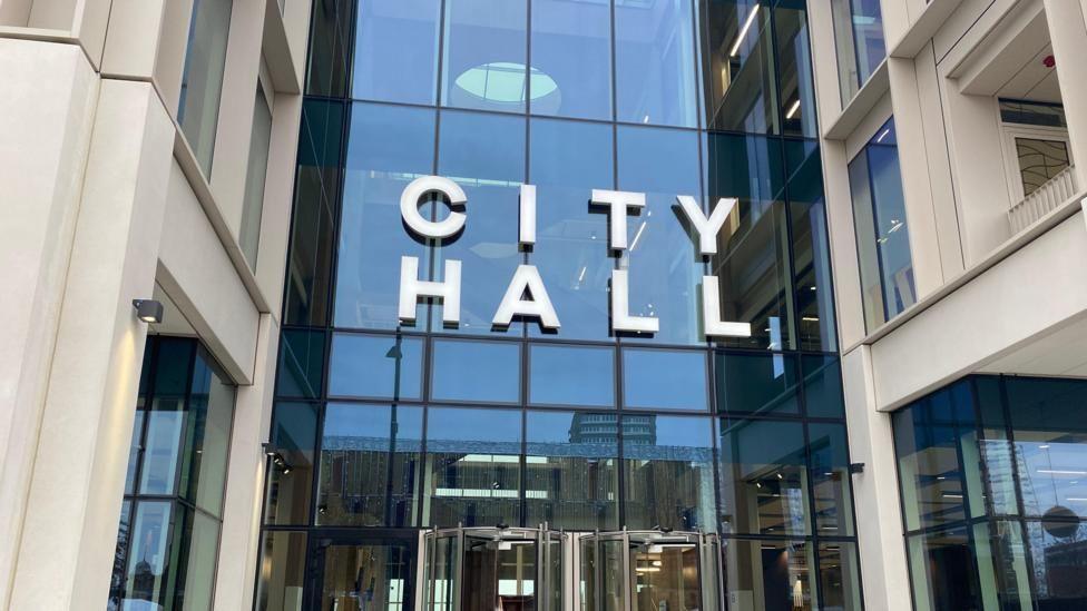 The image shows the entrance to Sunderland City Council. It's a modern building with the words City Hall above a glass fronted entrance and revolving doors 