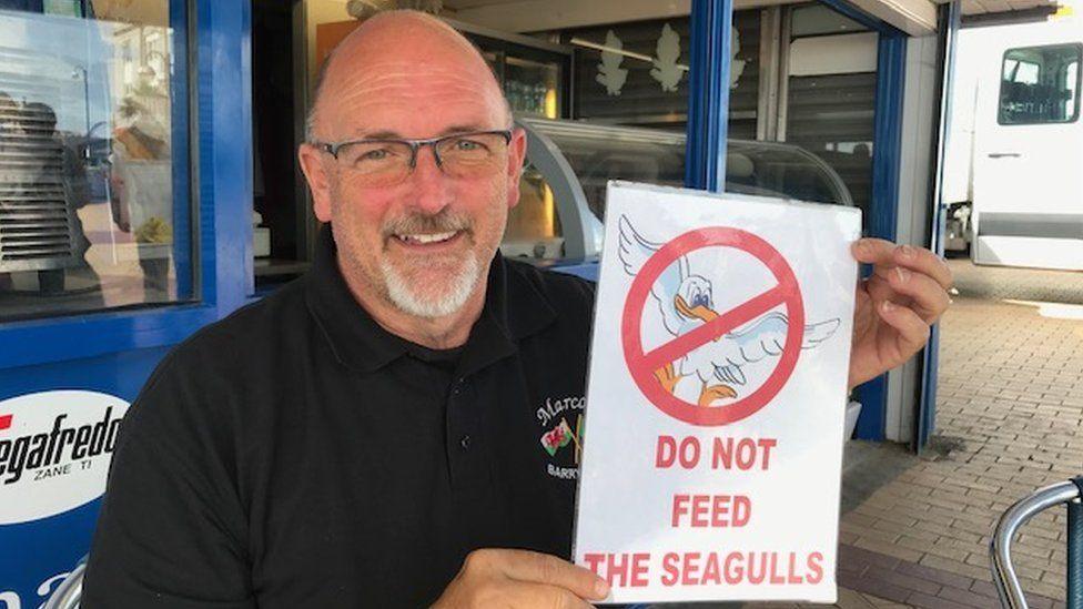 Marco Zeraschi outside Marco's cafe in 2019, smiling at the camera and holding up a sign reading 'Do not feed the seagulls'