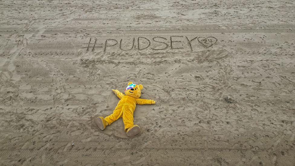 Pudsey Bears lies on a beach with his arms outstretched with the Pudsey 24 hashtag written in the sand above his head