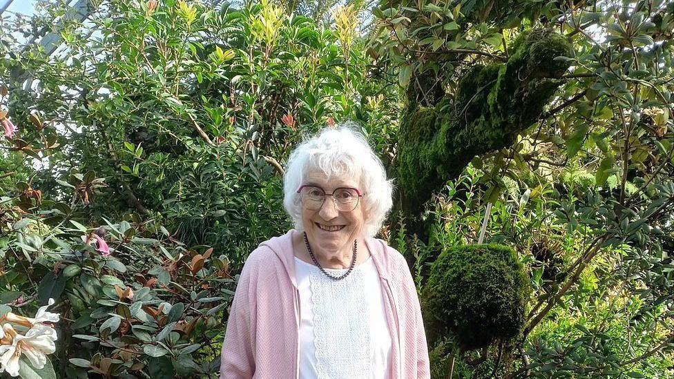 Maureen is wearing a pink cardigan over a white top. She has beige trousers on a a brown necklace. She is standing in front of bushes inside a hot house. She is smiling.