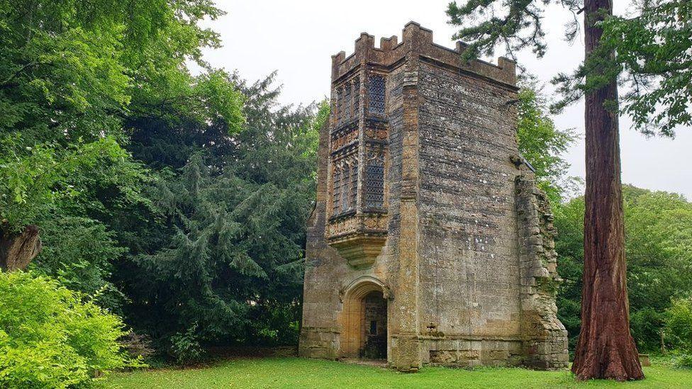 A crenelated stone tower with a large arched doorway. Above the door are two ornate bay windows, one above the other. The tower has broken stones on one side suggesting it was part of a larger building. It is surrounded by trees.