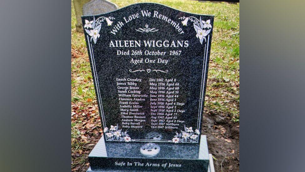 The gravestone inn the cemetery showing all the names of the babies, children and adults that are buried there. It is black marble with flowers in each corner. Aileen Wiggans name is at the top and the base says 'safe in the arms of Jesus'. 