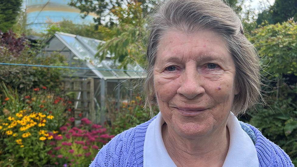 Smiling Pat Longton standing in her garden surrounded by flowers and trees with a greenhouse in the background