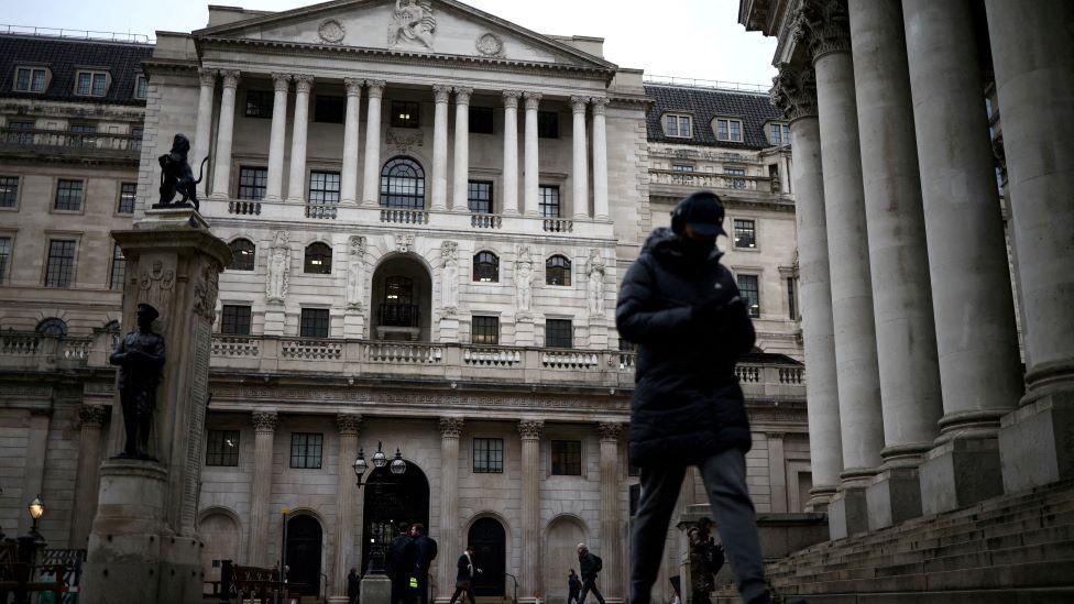 The exterior of a grand stone building - the Bank of England. A man in a long black coat, grey trousers and a black cap walks past
