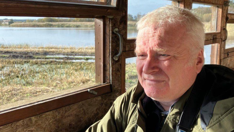 Minsmere site manager Nick Forster looks away from the camera while sitting in an observation building at the nature reserve. He has short grey hair and wears a green coat.