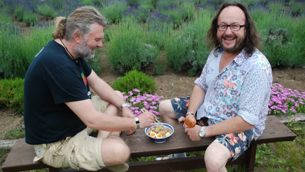 Si King and Dave Myers sitting and laughing on a bench with a bowl of food