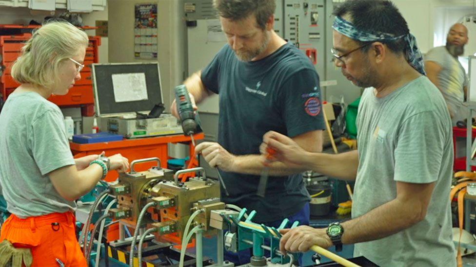 Three members of Léon Thévenin staff - a woman wearing orange trousers and a grey T-shirt, a man wearing a grey T-shirt and a bandana and another man in a navy T-shirt holding a drill. They are fixing a cable.