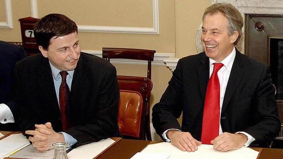 Douglas Alexander in a dark suit with a blue shirt and maroon tie looks and smiles at Tony Blair who is laughing and dressed in a dark jacket, white shirt and red tie. They are sitting at a table with papers on it. 