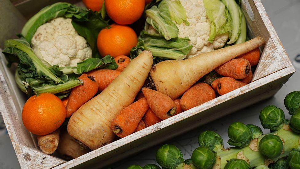 A crate of fresh fruit and vegetables, including carrots, cauliflower, parsnips, brussel sprouts, apples, and oranges