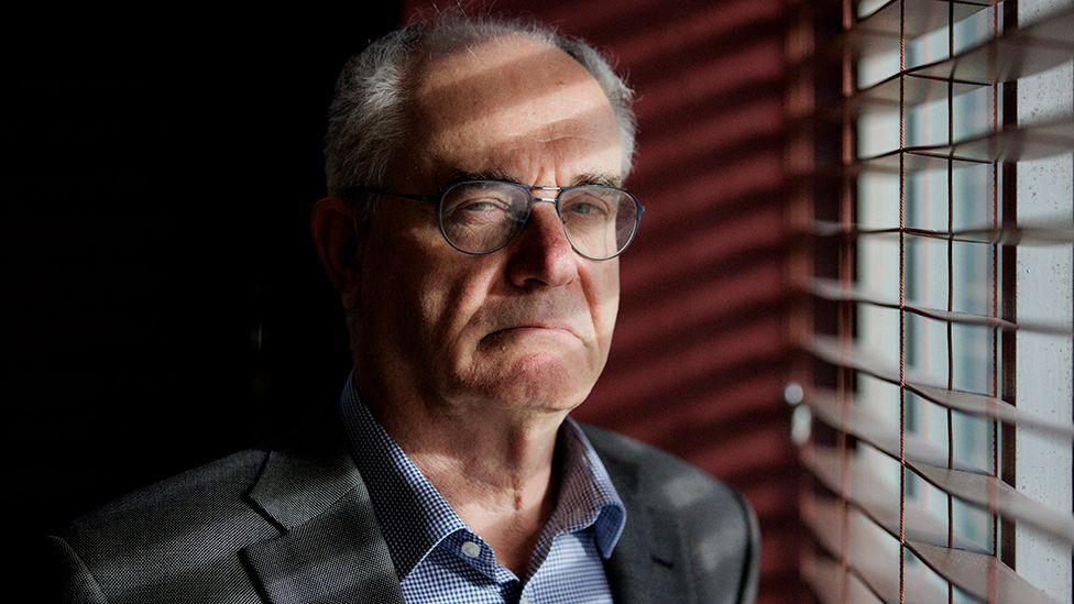 Clive Ruggles, who is looking serious and not smiling, is standing next to a window with wooden slatted blinds. He is wearing glasses and a grey suit with blue check shirt and has short white grey hair.