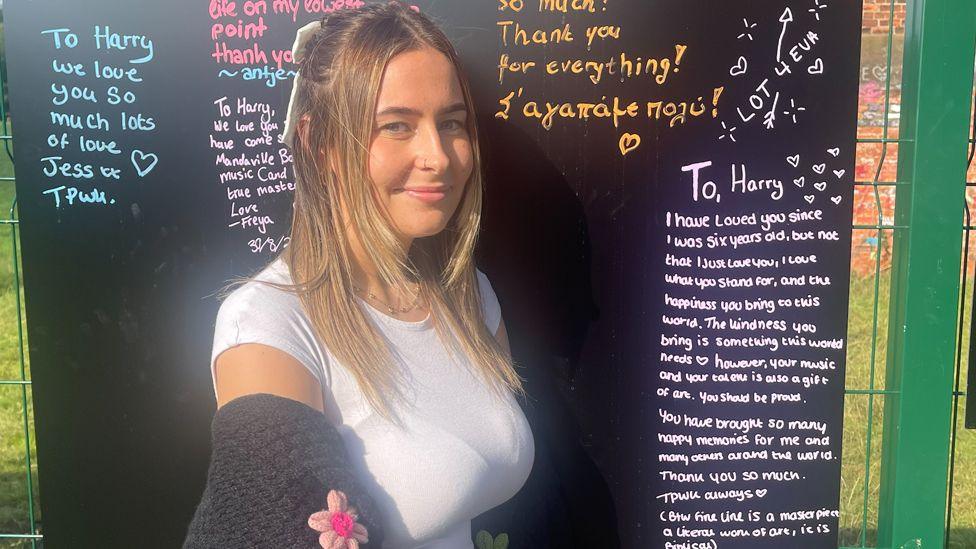 A woman with long hair is dressed in a white top with and woollen flowery cardigan wrapped around her arms. She stands in front of a green fence panel with a chalkboard fastened to it. The chalkboard has various messages addressed to Harry Styles