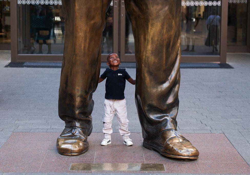 A boy in a black t-shirt and white trousers and trainers, looks up and smiles. He is standing between the feet of a statue. The glass entrance doors of a building can be seen in the background