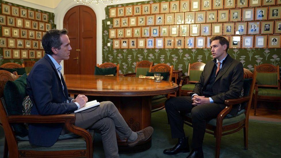 Mark Lowen sits in the Nobel Prize committee room around a wooden circular table with chairman Jorgen Watne Frydnes. Both are dressed in suit jackets, ties and trousers. In the background there are photographs on the wall of all the Nobel Peace Prize winners in frames.