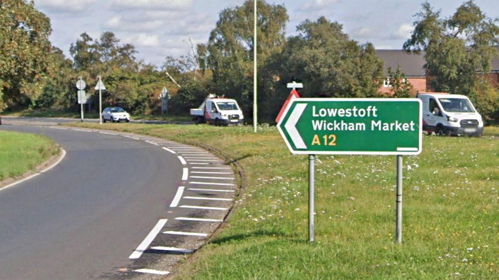 A green road sign for the A12 at Woodbridge, pointing in the direction of Lowestoft and Wickham Market. It is positioned on the verge of a sliproad off a roundabout.