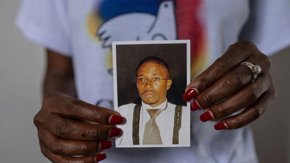 A close-up of a woman's hands, with read nail varnish, holding up a small photo of Floribert Bwana Chui Bin Kositi. In it he is staring ahead and looking serious. He is wearing oval wire-rimmed glassed, a silver and black dog tooth design tie, black braces and a white shirt. The woman's T-shirt, showing a peace dove design, can be seen in the background.