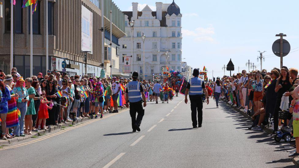 People line the streets of Brighton for the Brighton Pride parade. There are two police officers in the middle of the road, and hundreds either side of it. 