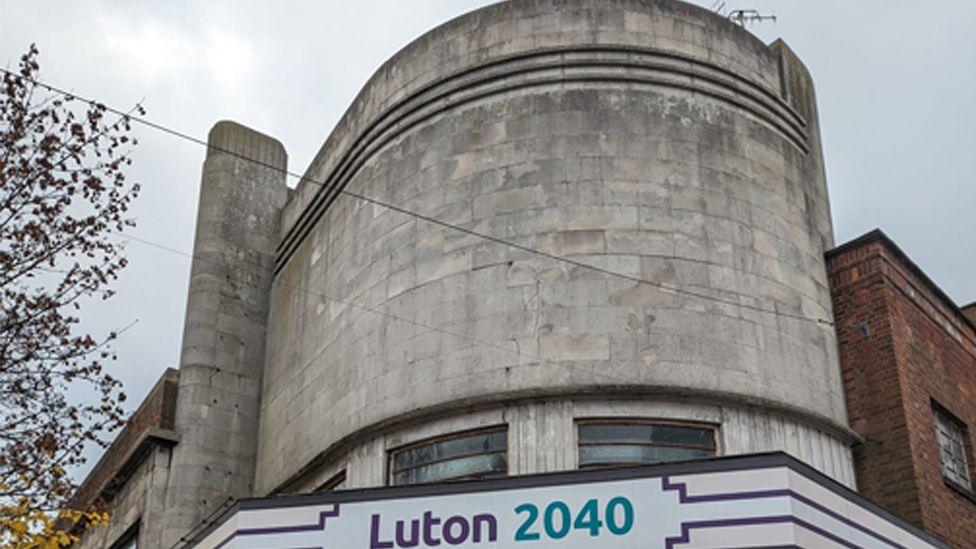 An empty Art Deco cinema in Luton. It shows a building, with light curved brick, with a board with Luton 2040 written on it.  There are windows above and two main doors to the building. There are signs around the front door.