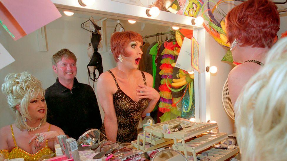 Basil Newby looking in a dressing room mirror in between two drag artists