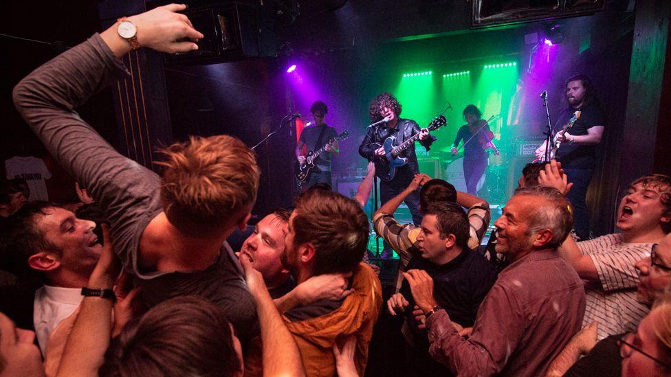 A man in a grey jumper jumps backwards with his hand in the air into a crowd of men, who are dancing in front of a green and pink-lit stage where a band of three guitarists and a violin player, all dressed in black and standing in front of microphones, play their music