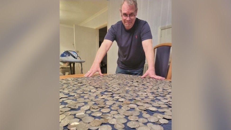 Steve Hickman, a man stood over a wooden table. On the table is a blue tablecloth which has a hoard of coins on top of it. He is wearing a blue T-shirt and pair of glasses and is looking over the hoard towards the camera. 