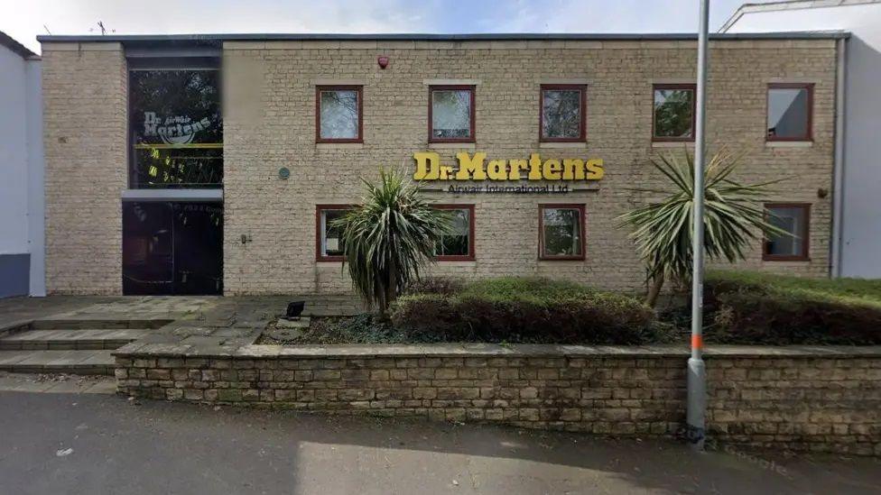 Two-storey brick-built factory with small square windows. The words Dr Martens are fixed to the wall in yellow letters. There are steps to the left and a low wall in front of the factory.