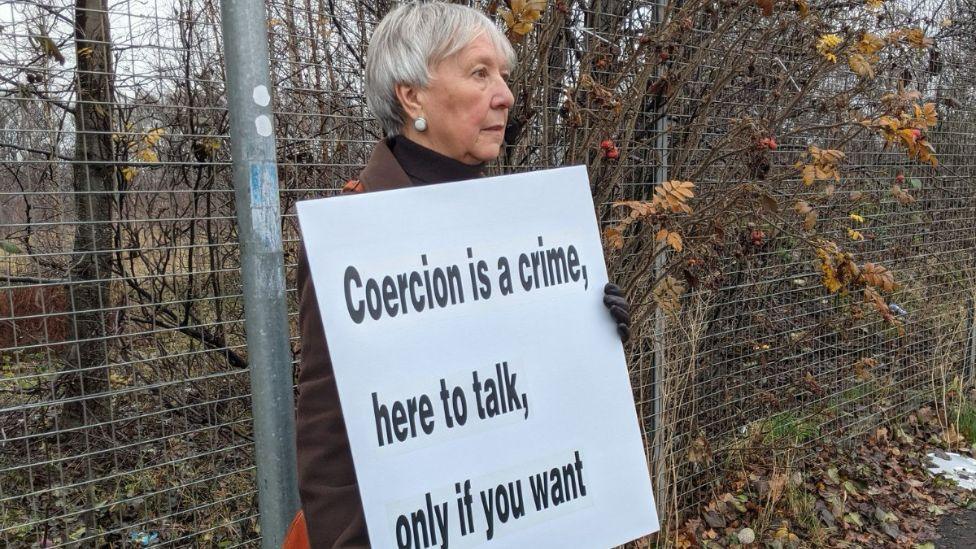 A woman standing outside in front of a metal wire fence holds a white sign saying "coercion is a crime, here to talk, only if you want". She has short grey hair and is wearing a brown jacket and gloves. 
