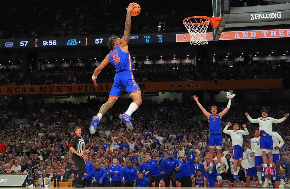 Basketball player Alijah Martin leaps high toward the hoop for a slam dunk during a game. The player wears a blue and orange uniform, and team-mates on the bench celebrate in the background as a packed arena watches.