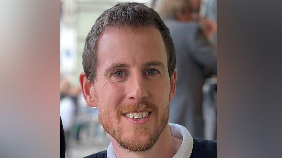 Man with short brown hair and beard smiling at the camera. He is wearing a blue sweater with a white collar