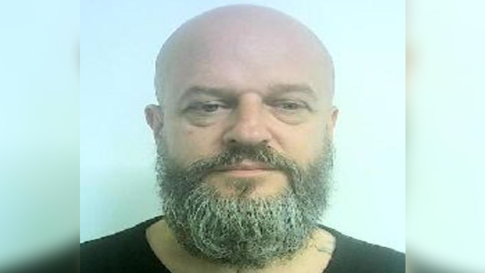 A bald man with a grey/ brown beard standing in front of a blue background wearing a black t-shirt.