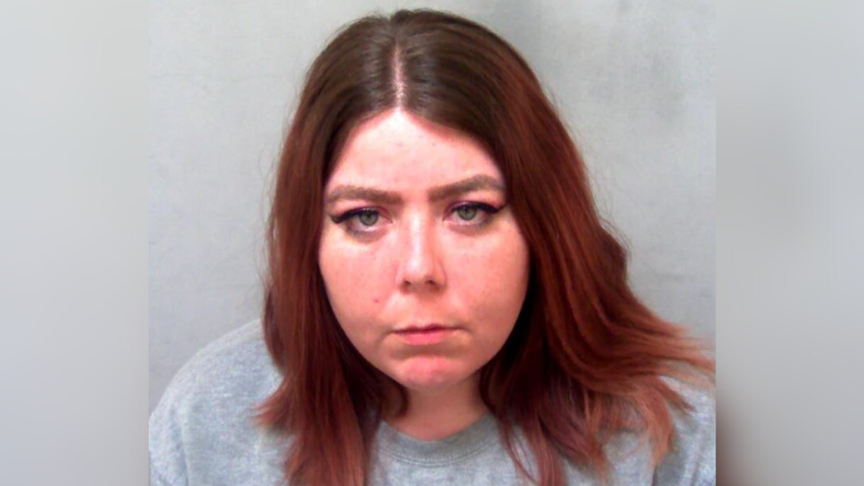 A custody mugshot of Lucinda Fyfield. She has long red hair and is wearing a grey sweater. She is looking at the camera with a slightly angry expression.