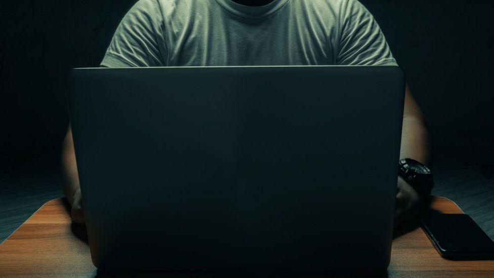 A man sits in front of a laptop computer, we see only his toros and not his face. a mobile phone sits beside the laptop