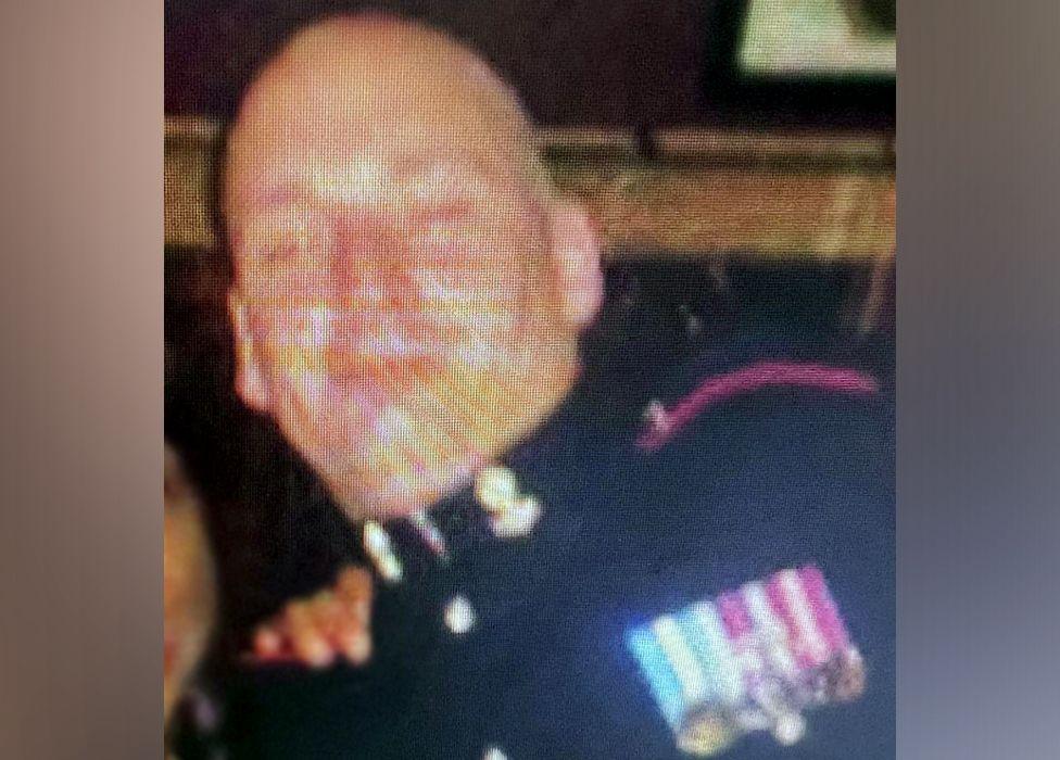 A blurred image of Michael Webber - he's wearing a dark-coloured uniform and has medals on the left side with ribbons of purple/white and blue/white. He's bald.