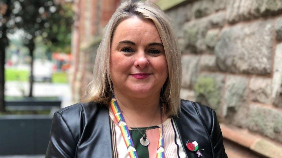 Sandra Duffy stands outside Derry's Guildhall. She has blond hair and is smiling slightly. Her black leather jacket is being worn over a many coloured top