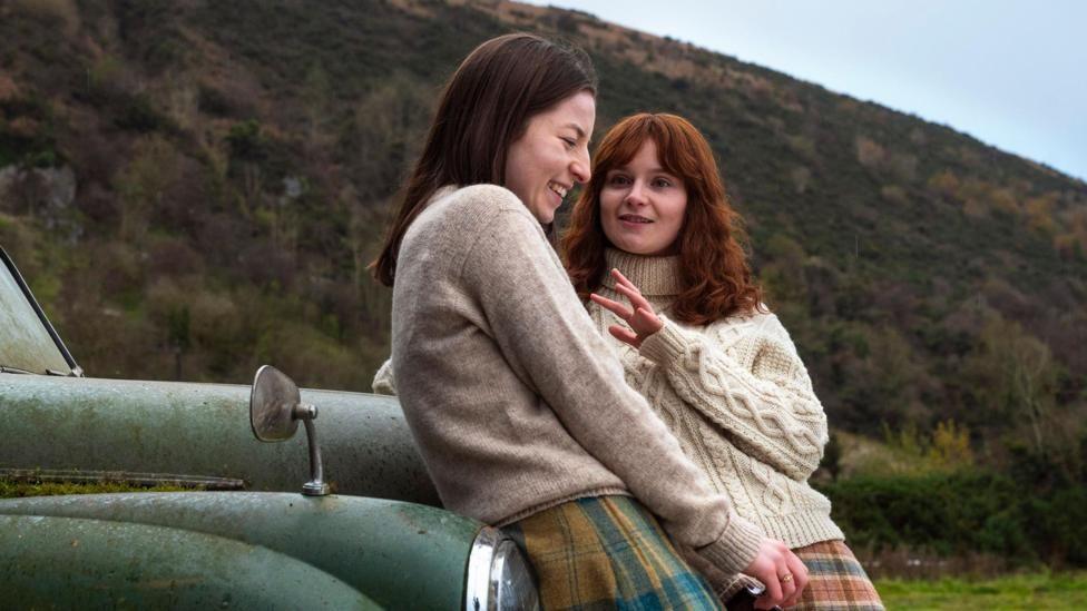 Hazel Doupe and Lola Petticrew in character, they are both leaning against an old green car, there is a mountain in the background. Hazel Doupe is wearing a cream jumper with a yellow and blue skirt. Lola Petticrew is wearing a light cream jumper with a red, yellow and blue skirt
