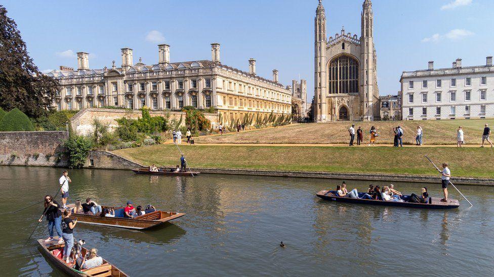 Four punts on the River Cam along the Backs on a summer's day. Each one has one or two people standing up and steering the flat-bottomed boats. Beyond the river is a shorn meadow running up to King's College Chapel.