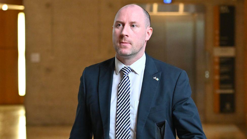 Neil Gray, a bald man wearing a dark suit, white shirt and stripy tie, in a medium close-up shot in the Scottish Parliament. 