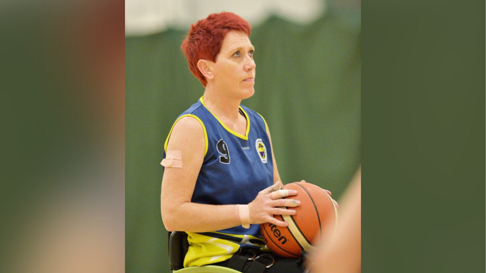 Ms Carey playing wheelchair basketball. She wears a blue vest with a yellow design on the arms and sides. It has a number nine in the top left corner and a blue and yellow circle logo in the top right. Ms Carey has the basketball in her hands and is looking towards the direction of the net, away from the camera, with a look of concentration on her face. She has short red hair and plasters on her right upper arm, wrist, and on some fingers. 