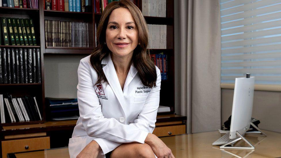 Dr Hayley Brown is sitting on a desk wearing white doctor's coat. She is smiling and has long brown hair.