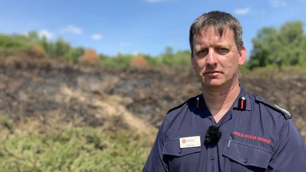 Andy Cole pictured in blue firefighter's uniform shirt in front of an area of burnt heathland during an incident in 2023