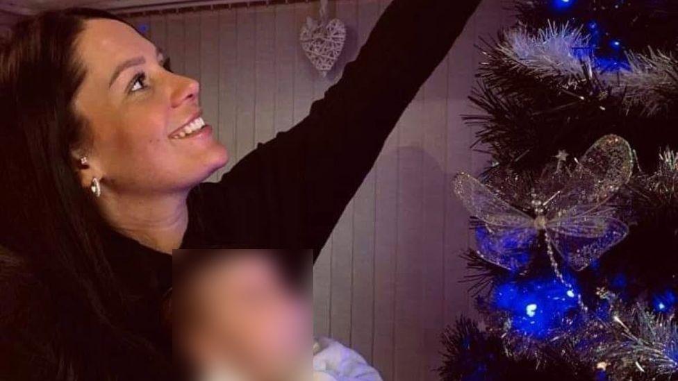 A smiling Kiena Dawes, who has long black hair, reaches up to the top of a Christmas tree while holding a baby girl in her left arm.