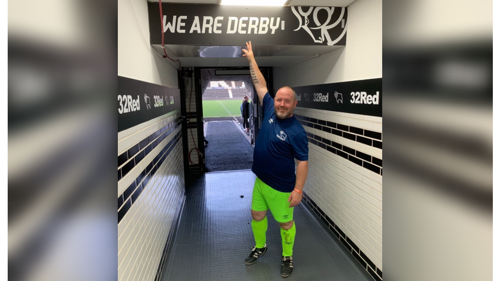 Bald man with navy football shirt and neon green football shorts and socks pointing to a Derby County Football Club sign saying 'we are Derby' in a corridor leading to a football pitch in the background
