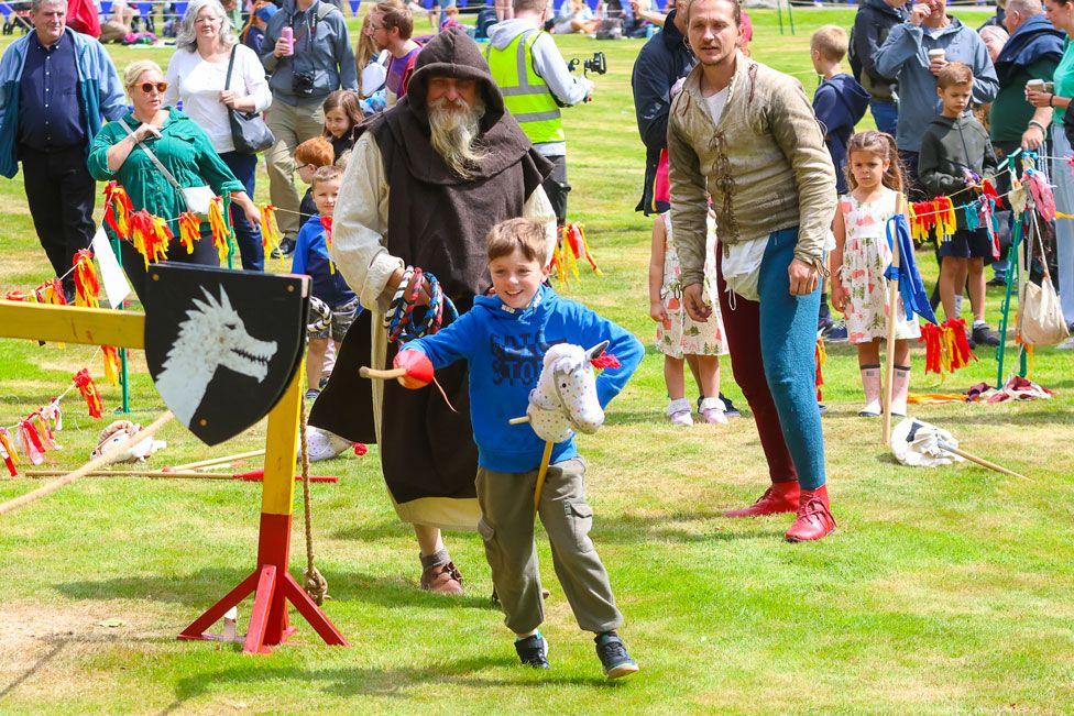 A young boy on a pretend horse charges with a small wooden lance while group of people look on in the background