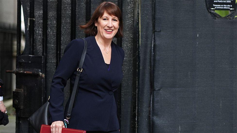 Chancellor Rachel Reeves in a dark blue dress carrying a folder. She has short dark hair and is walking in front of a black gate