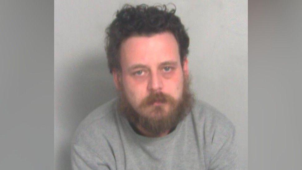 Close-up image of Joshua Robinson. He has dark brown short wavy hair and a full beard. He is wearing a grey jumper.