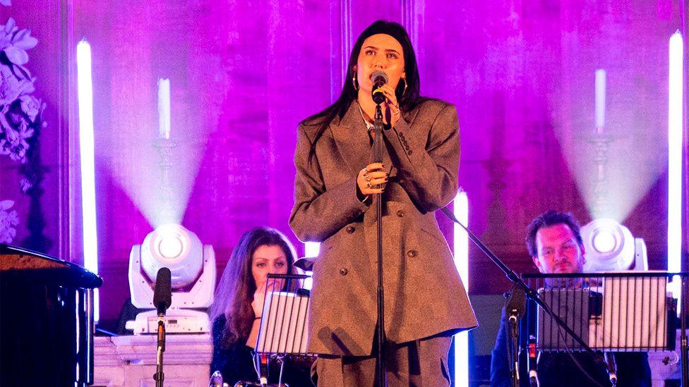 Sienna Spiro on stage in London, wearing a blazer/suit, with a band ons tage with her. There are bright spotlights behind her and a purple haze.