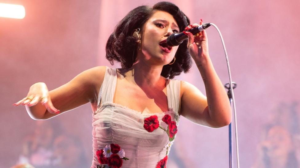 Singer RAYE, in a white dress embroidered with flowers, sings into a microphone