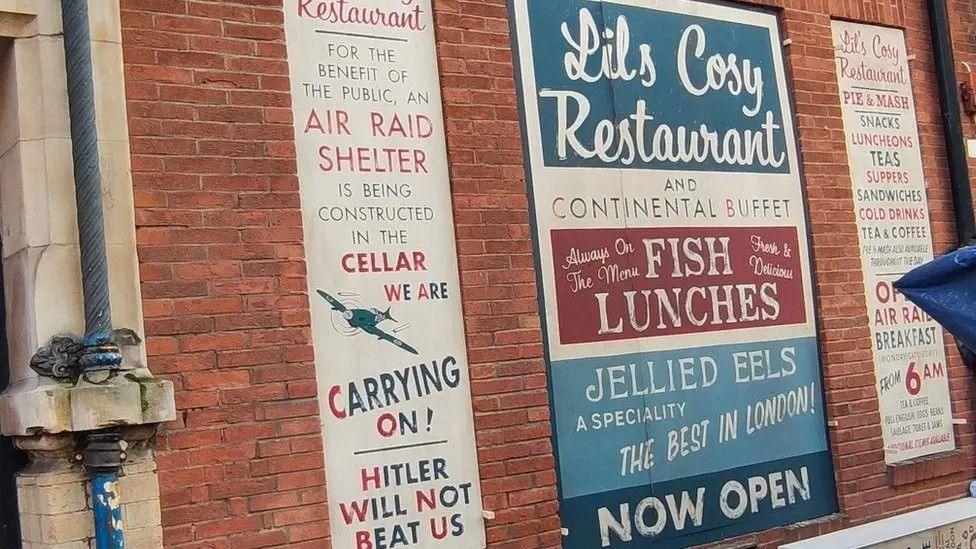 Mocked up posters on a wall - one says air raid shelter being constructed in cellar - "We are carrying on - Hitler will not defeat us". Another advertises jellied eels.