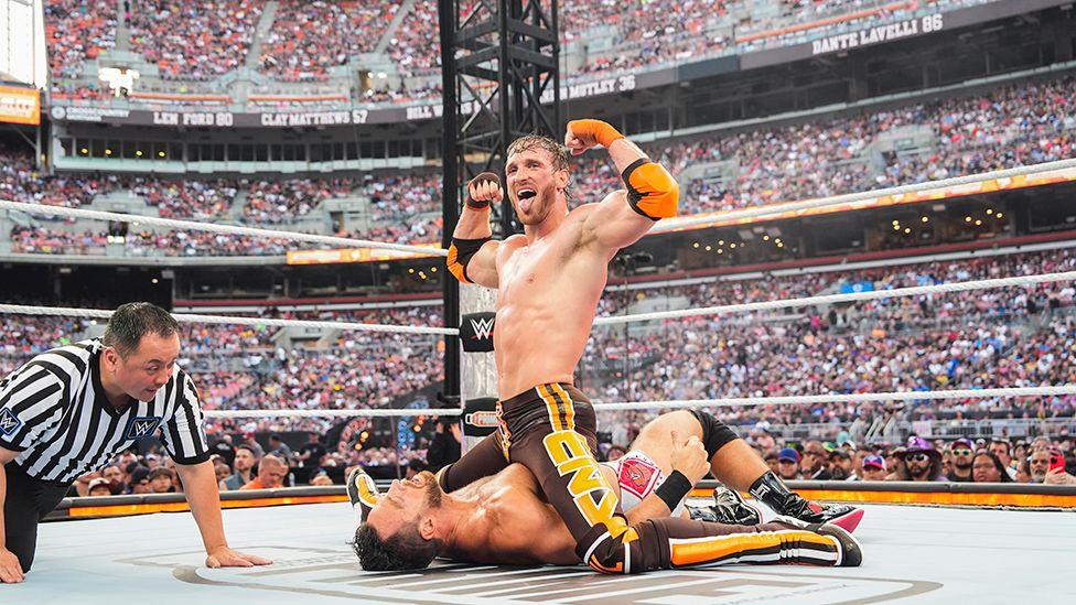 Picture of a ring at a wrestling match. In the middle, Logan Paul has trapped his opponent on the floor and is astride him, and making a triumphant gesture to what looks like a massive arena audience. He is holding his arms aloft and sticking out his tongue. Next to them, a referee is on his hands and knees, monitoring the wrestling match.