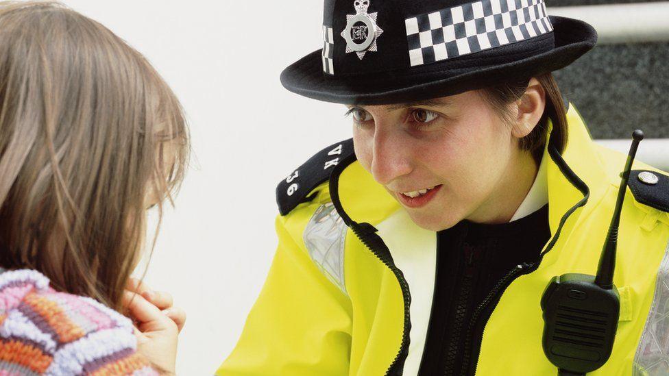 Female police officer in uniform knelt down speaking to a young girl who has her back to the camera